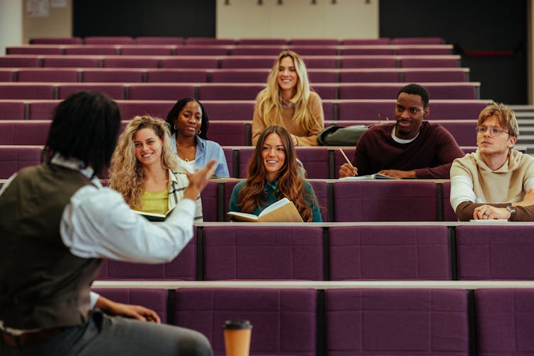 A young Black man is sitting in front of students in a lecture hall, gesturing as they smile