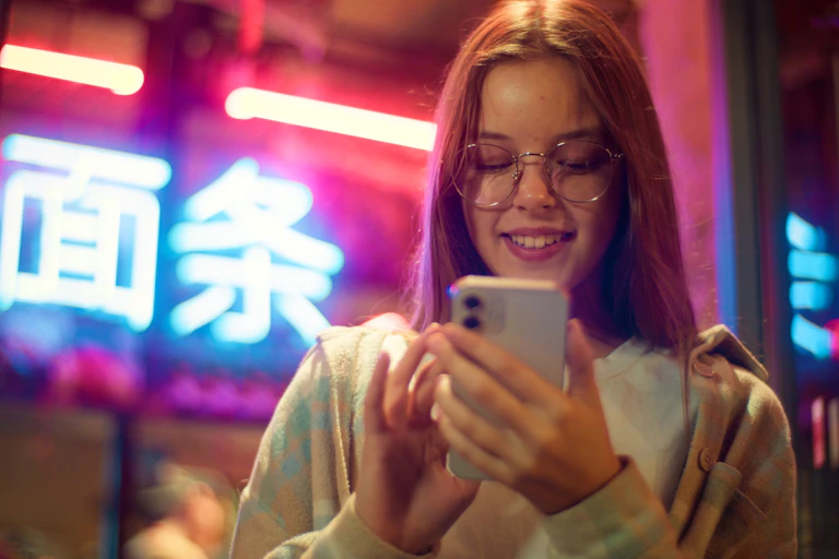 A smiling teenager looking at their phone in a neon city.