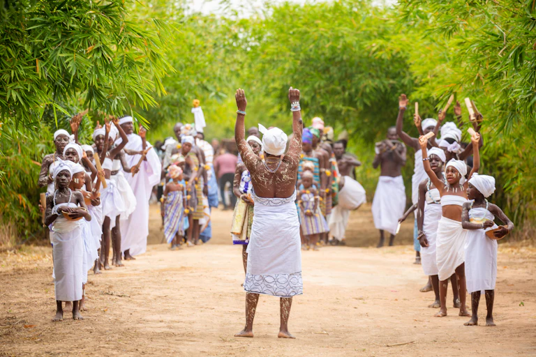 A group of women and girls on a dirt road, dressed in white traditional garments. They stand in rows, some raising their arms.