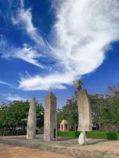 Three very tall sculptures, one a stylised human figure the other two buildings, against a backdrop of leafy trees.