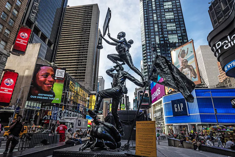 A sculpture of three human figures in motion balanced on top of one another in the centre of a bustling urban space with billboards and skyscrapers.