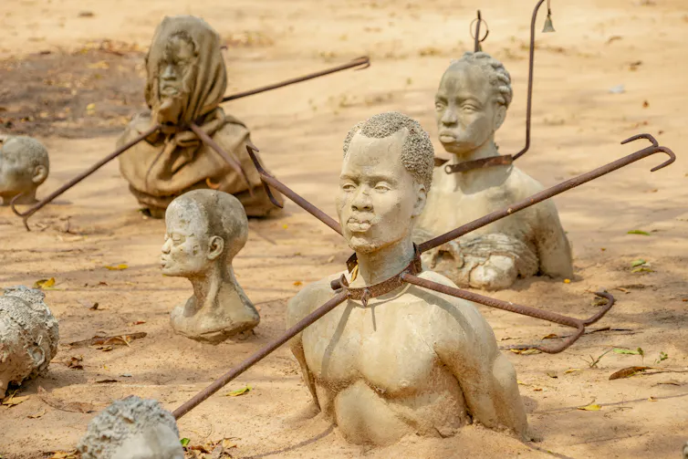 Sculptures of human heads and torsos emerging from the earth, metal collars around their necks.