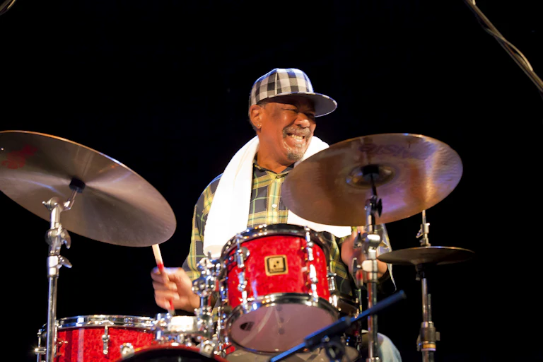 An older man smiles broadly as he sits playing on a drum kit of bright red drums.