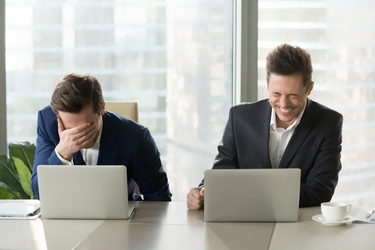 Two men in suits sit in front of laptops. The man on the left covers his face in embarrassment while the man on the right laughs.