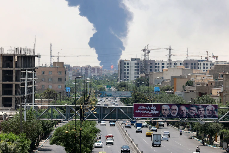 A dark plume of smoke is seen in the dispace as as billboard with the faces of several men is seen in the foreground