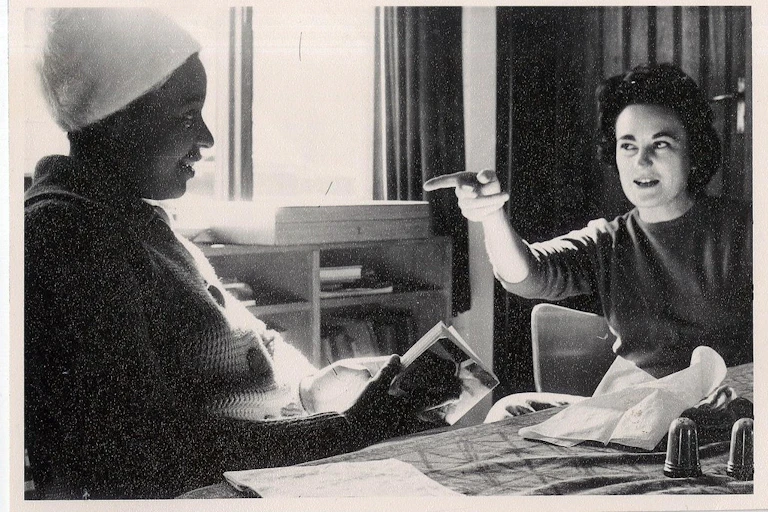 An elegant woman sits at a table and, with a challenging half smile, points a finger at an African woman in a headpiece sitting with her.