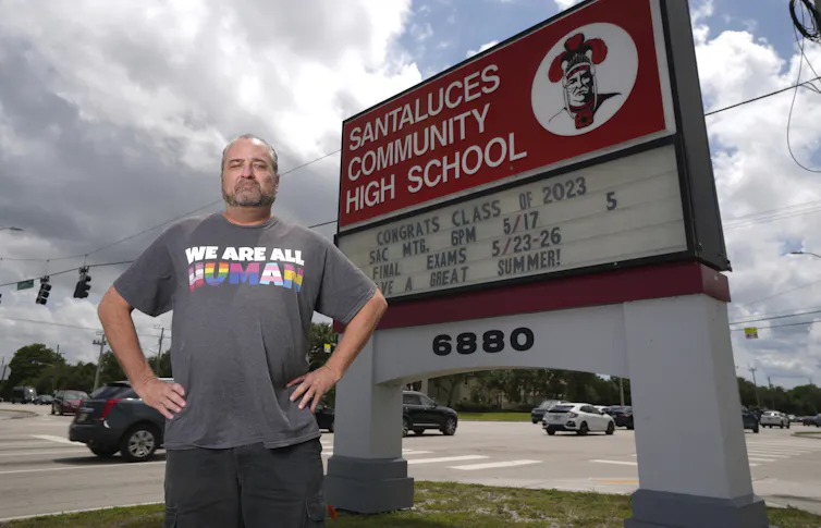 A person in a shirt that says 'we are all human' in front of a school sign.
