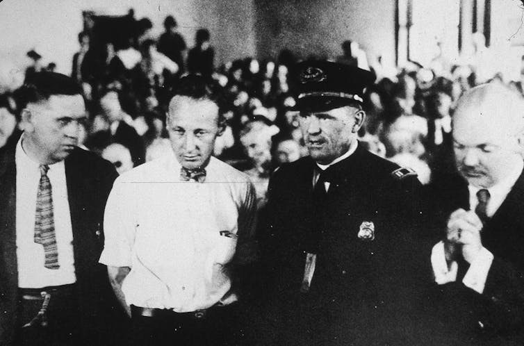 A black and white photograph of four men standing amid a crowded room, including one younger man in a white, short-sleeve shirt and bow tie.