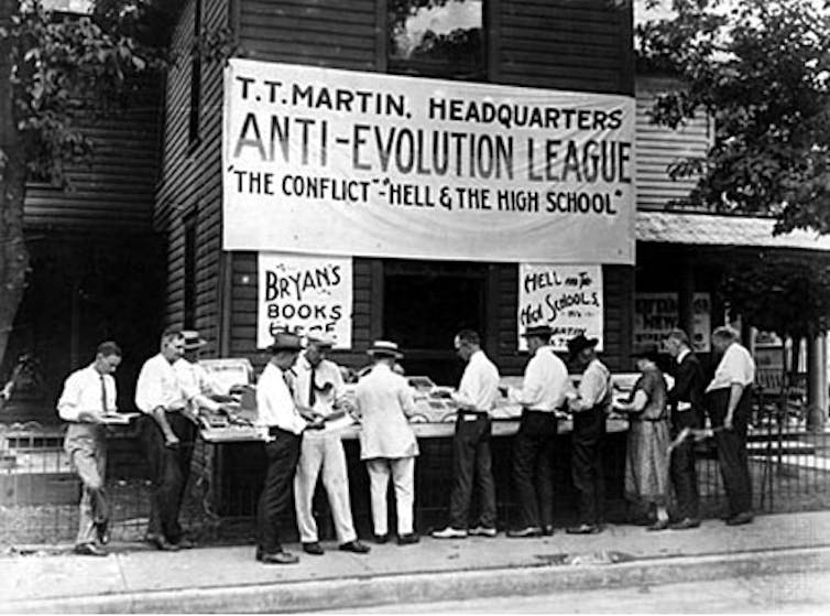 A black and white photo shows men in white shirts gathered around an outdoor book stall.
