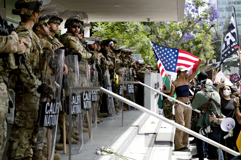 Protesters, one waving an American flag, stand face-to-face with heavily armed soldiers at the top of a staircase into a federal building.