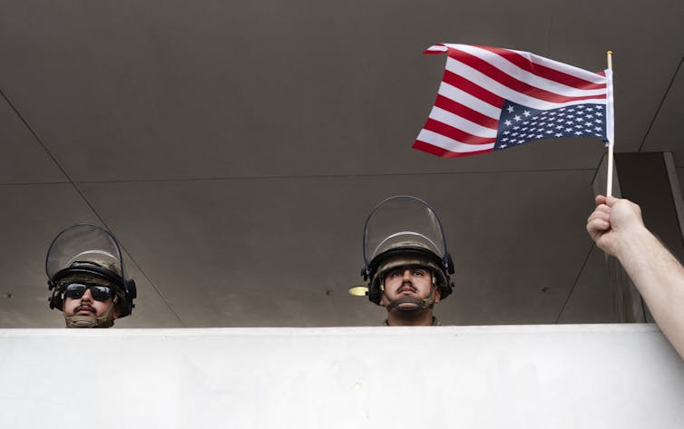 The heads of two National Guards in helmets look over a retaining wall as someone waves an American flag in front of them.