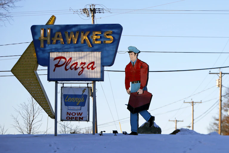 Large vintage sign reading "Hawkes Plaza" next to a large billboard of a man in a red jacket holding a briefcase.