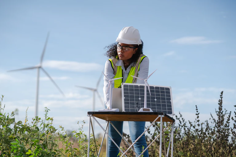 A woman stands outdoors near wind turbines, wearing a hard hat and typing on a solar powered laptop