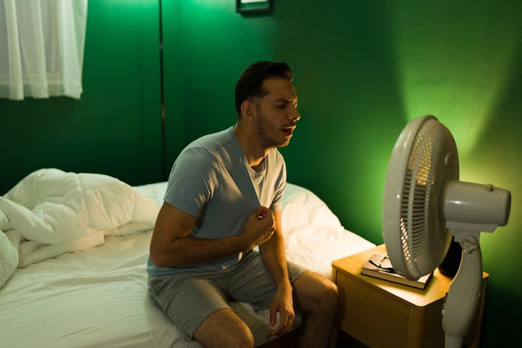 A man sits on his bed in front of a fan cooling himself off.