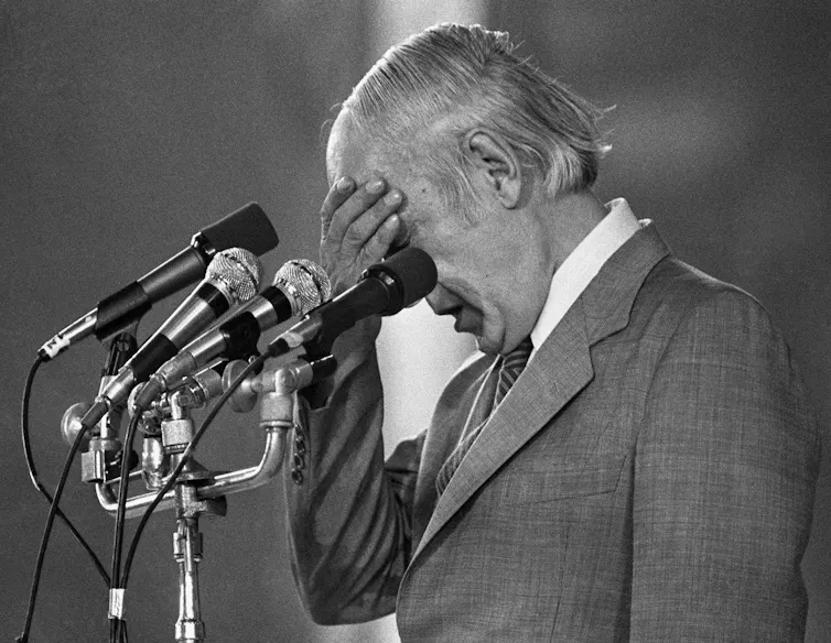 A man with thinning grey hair bows his head with his hand on his forehead behind a bank of microphones.