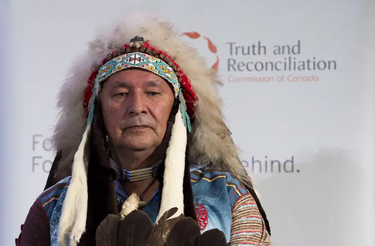 A man in an Indigenous head dress sits in front of a banner that reads Truth and Reconciliation