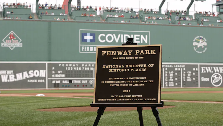 A plaque honoring Fenway Park is displayed on an easel on a baseball field.