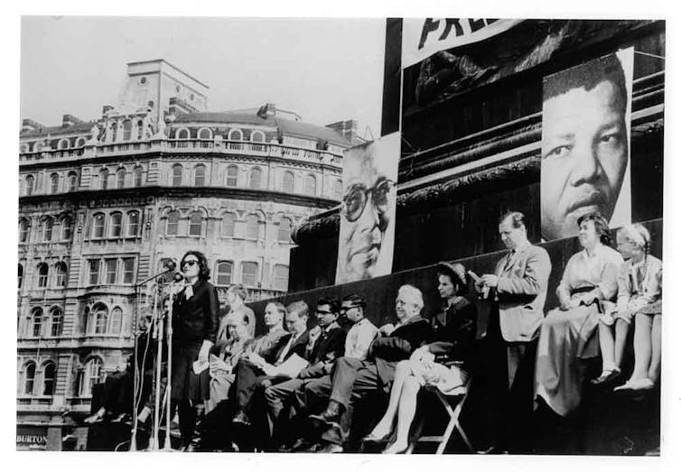 An old photograph of a woman on a stage speaking into a microphone with a row of people in chairs behind her and large posters of two men behind them.