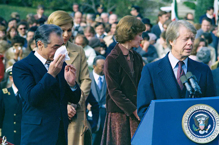 A man in a blue suit speaks in front of a lectern as two women and one man stand behind him.