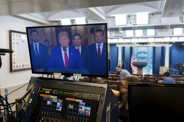 A man in a blue suit and red tie, speaking on a large monitor in a medium-sized room.