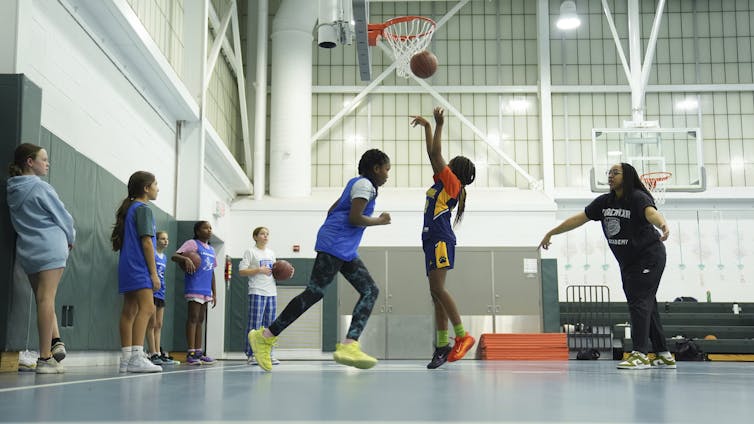 A young woman looks on as a young girl shoots a basketball in a hoop