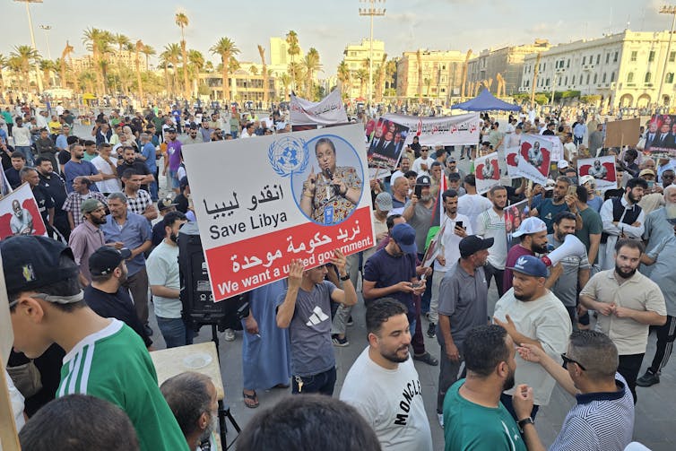 A large group of men stand in a crowd and hold up signs, with buildings seen in the background.