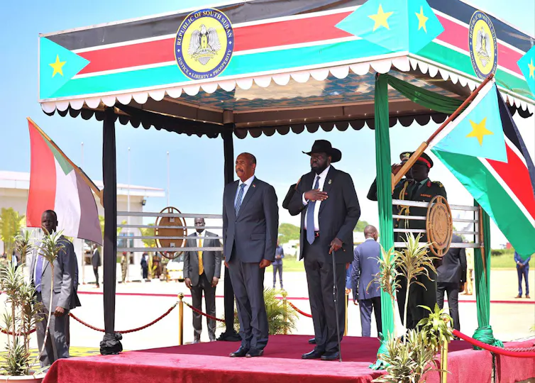 Two men in dark suits stand under a red, black and green canopy and look forward.