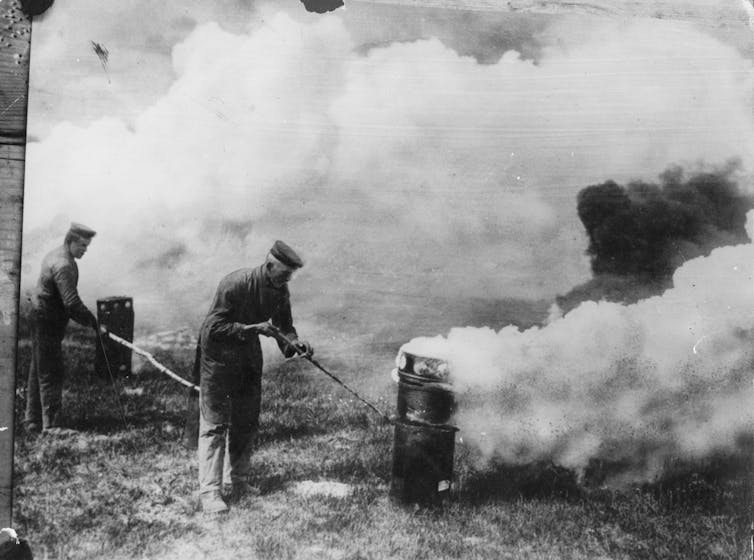 A vintage photograph of a soldier poking a cylinder, which releases a cloud of smoke.