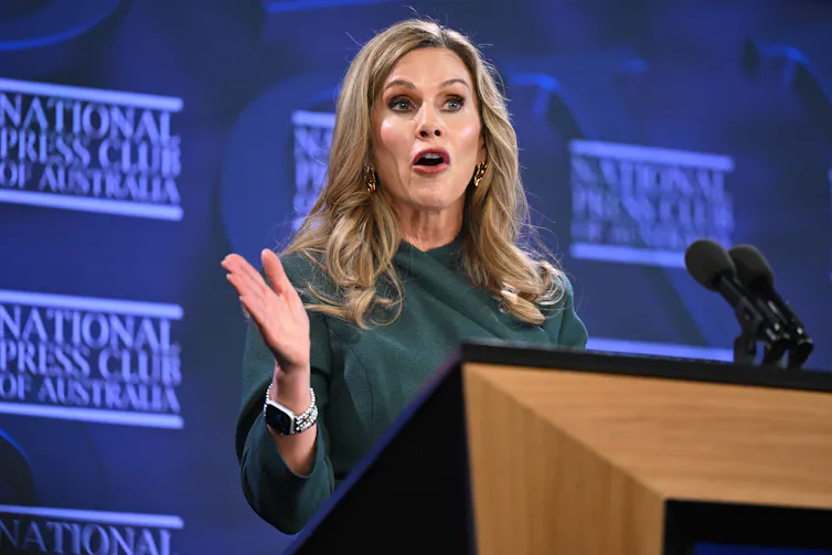 A woman with blonde hair wearing a green dress speaking on a stage.