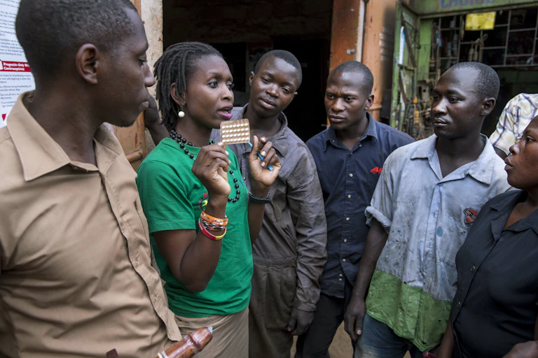 A woman talking to a group of men. She is holding a blister pack of pills.