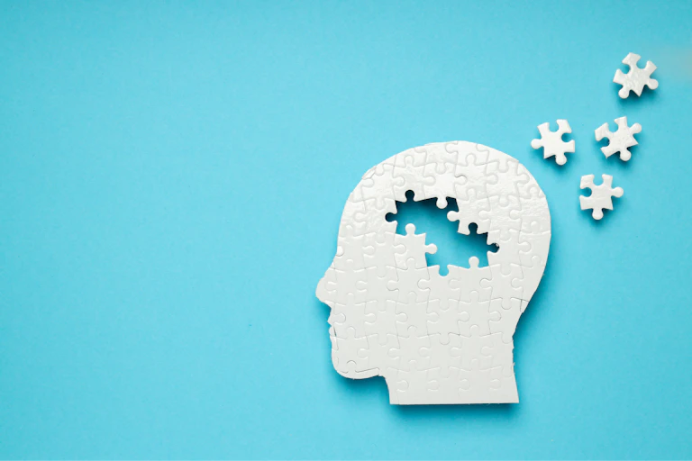 The silhouette of a white head on a blue background. There are puzzle pieces missing from the centre of the head, which are scattered on the blue background next to it.
