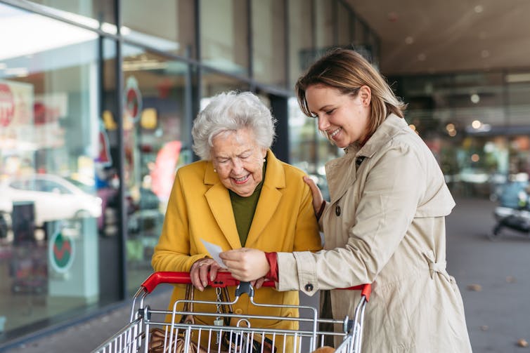 A middle aged daughter helps her older mother with shopping at a grocery store. The mother leans against a shopping cart while the two look at a shopping list together.