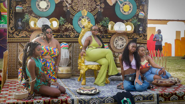 Five young African women in bright clothing pose for a photograph in a decorated area with colourful prints, a man standing off to the side in the distance.