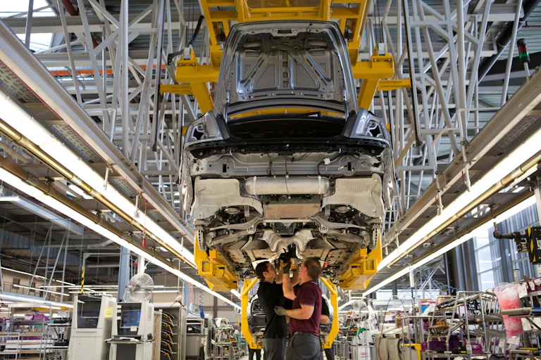 The body of a car suspended over a factory floor with workers attending its underside.