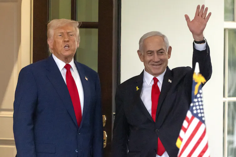 Two men in suits stand outside a white stone building, the shorter bald one waves while the rotund man with orange-blond hair grimaces.