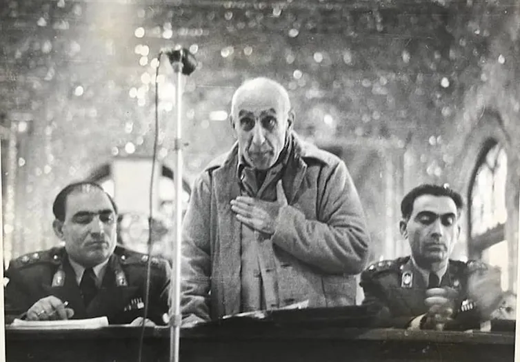 An elderly man holds his hand over his heart, flanked by men in military uniform.