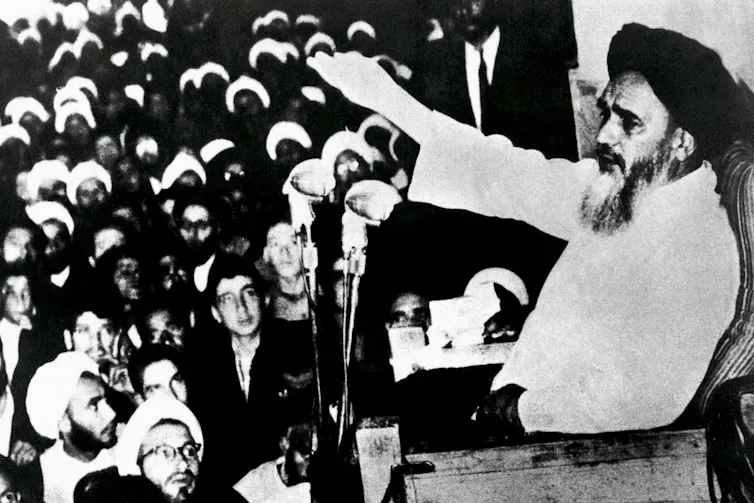 A middle-aged man with a beard in traditional Islamic clothing gestures to a crowd as he makes a speech.