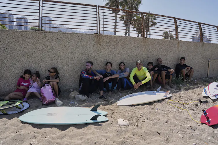 Surfers with surf boards crouch against a concrete retaining wall on a beach.