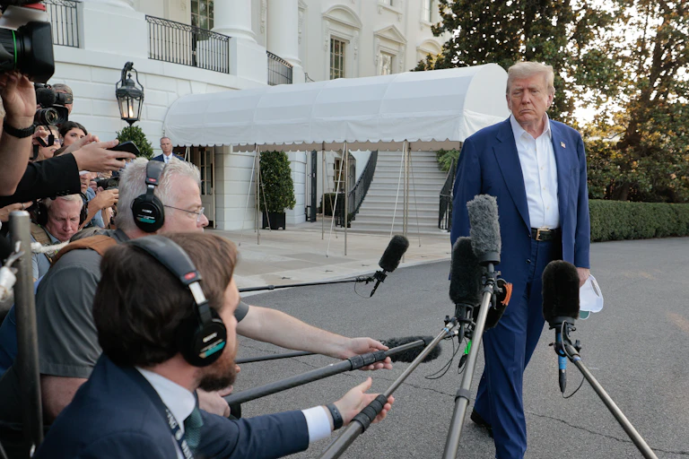 An older man with light hair and a blue suit and white shirt stands in front of a white building and in front of a large crowd of people holding microphones in his direction.
