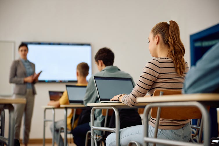 a teacher stands at the front of a class, students sit on desks using laptops