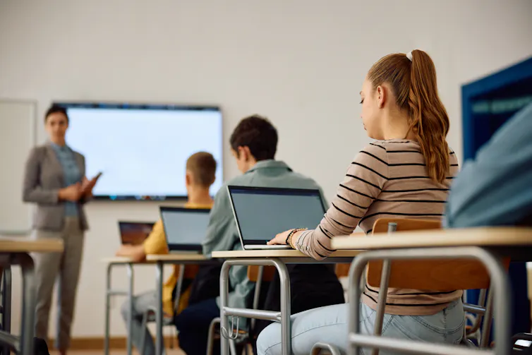 a teacher stands at the front of a class, students sit on desks using laptops