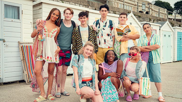 a group of teens stand in a family photo style - they are in front of beach changing cabins
