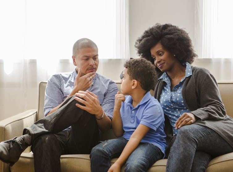 A Black couple sit on a couch, speaking with a young boy.