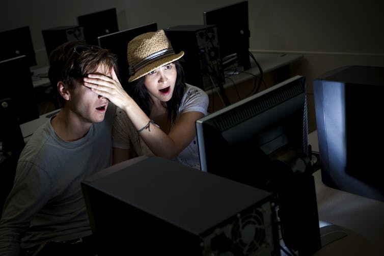 A girl covers the eyes of a boy while she looks at a computer screen in a darkened room.