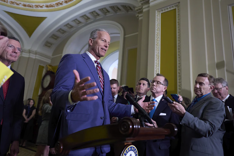 a white man in a blue suit gesticulates as he stands before microphones and a bunch of people holding recorders in an ornate building
