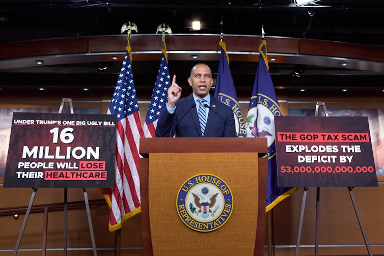 a Black man points the ceiling as he stands in front of a lectern and two poster boards