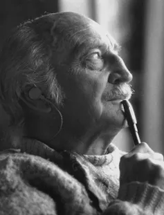 An elderly man with a white moustache and hair looks thoughtfully, a pen to his lip.