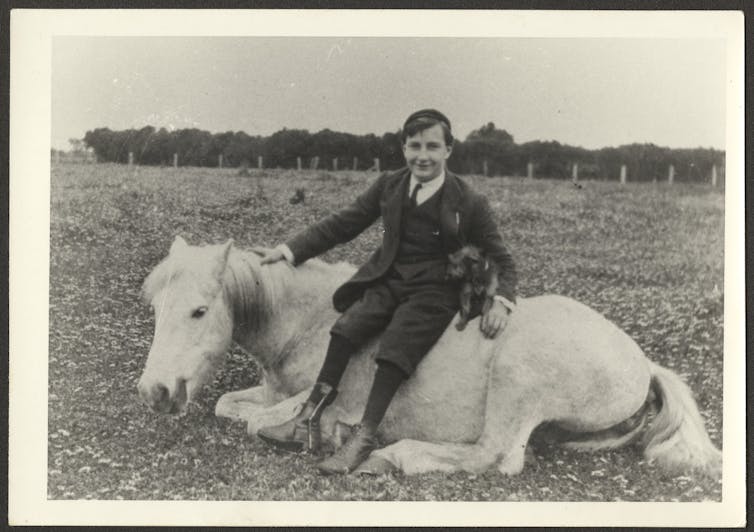Alan Marshall sitting on a horse, which lays beneath him in a field.