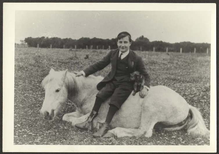 Alan Marshall sitting on a horse, which lays beneath him in a field.