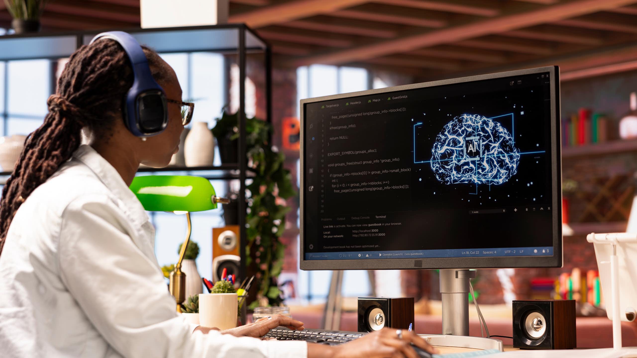 Mujer joven negra con gafas y auriculares mira una pantalla de ordenador con un cerebro digital dibujado con el símbolo de "AI" en el centro